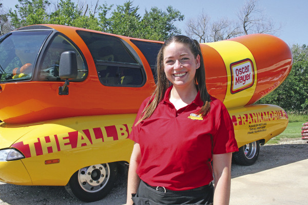 Chloe Van Caeseele poses in front of the Frankmobile during Saturday’s appearance at Hired Man’s Grocery &amp; Grill. The New York native is one of 12 young adults chosen this year to be Frankfurters – a position formerly known as a Hotdogger. They are representatives for Oscar Meyer. Travis Mounts/TSnews